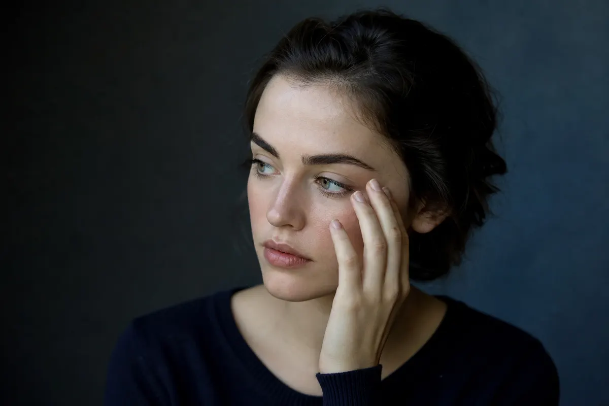 Portrait discret de trois quarts, main effleurant le contour de l'œil dans un geste de gêne légère, lumière naturelle douce, aucun texte ni objet visible.