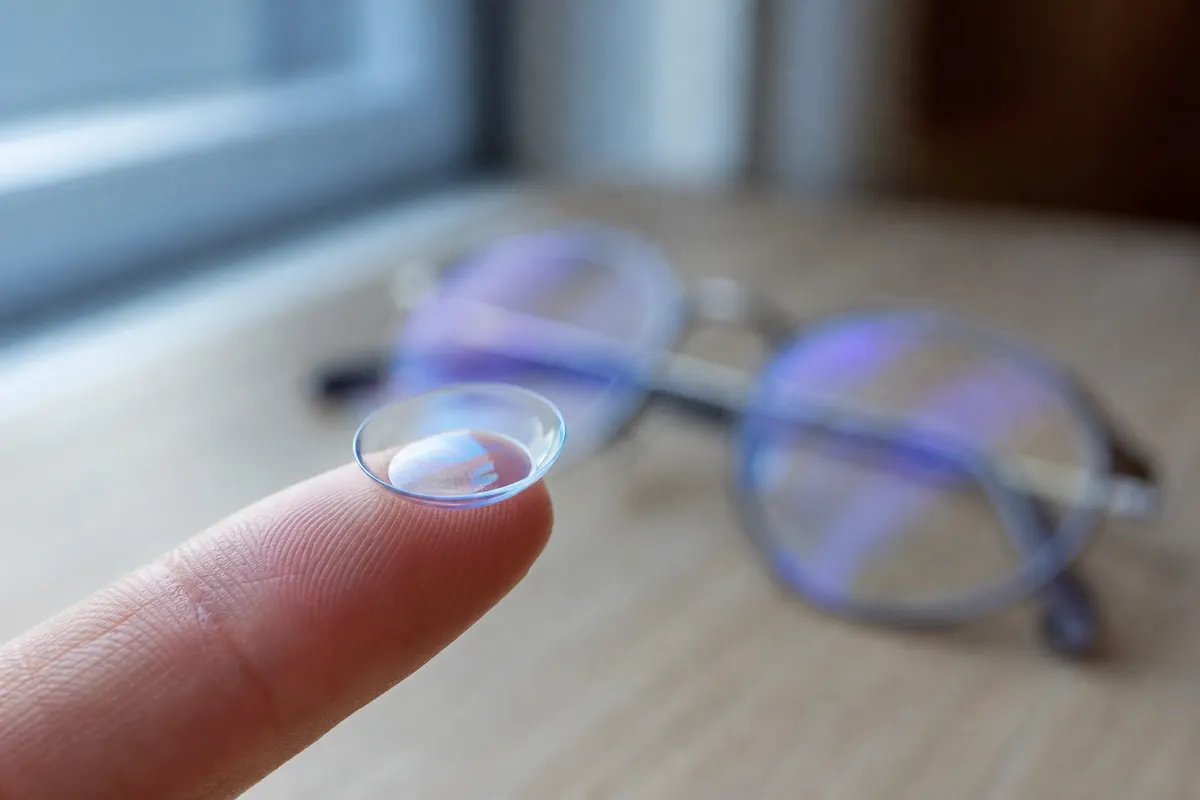 Gros plan sur une lentille de contact souple posée sur le bout d'un doigt, avec en arrière-plan flou une paire de lunettes de vue posée sur une table.
