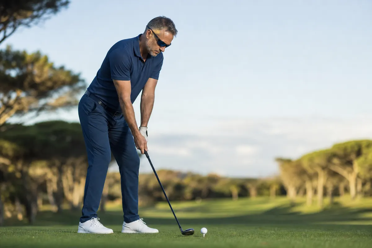 Golfeur aux cheveux grisonnants, lunettes de soleil enveloppantes, en position d'adresse au-dessus d'une balle blanche posée sur un tee, sur un parcours bordé de pins.