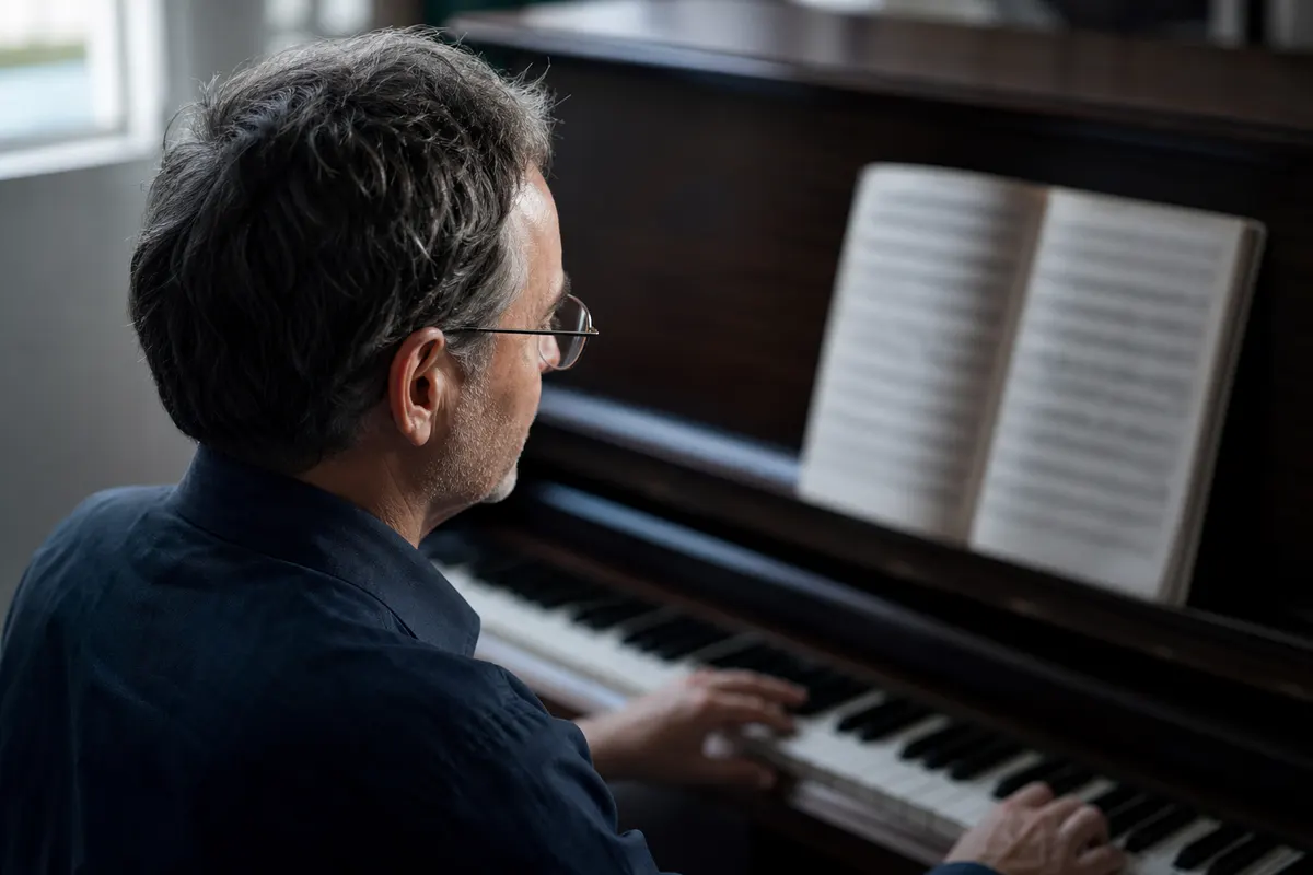 Musicien aux cheveux grisonnants, lunettes à monture fine, lisant une partition sur un pupitre derrière un piano droit, dans une pièce éclairée par une fenêtre.