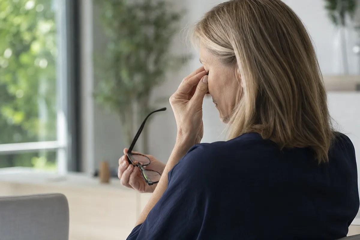 Femme d'une cinquantaine d'années retirant ses lunettes de vue pour se frotter doucement les yeux, lumière naturelle douce, aucun décor visible.