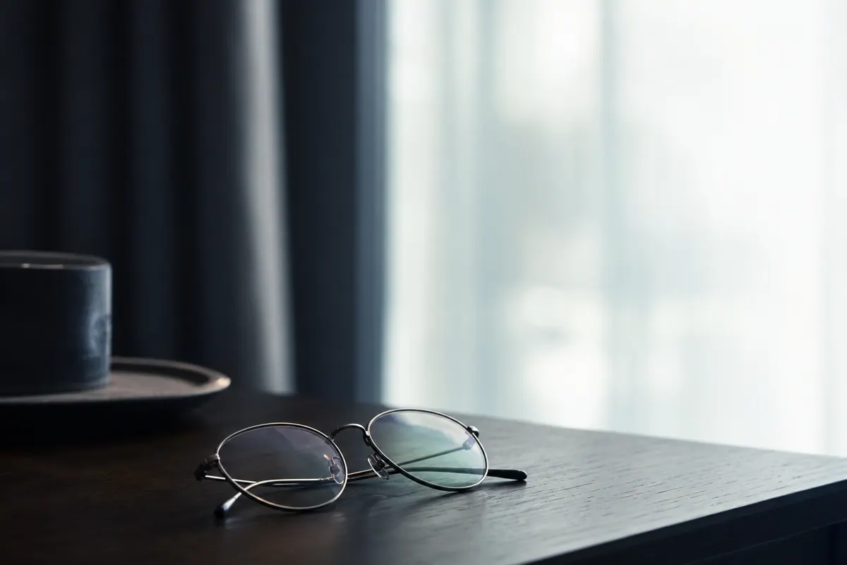 Paire de lunettes de vue posée sur une table près d’une fenêtre voilée, lumière tamisée et diffuse, ambiance calme et reposante.