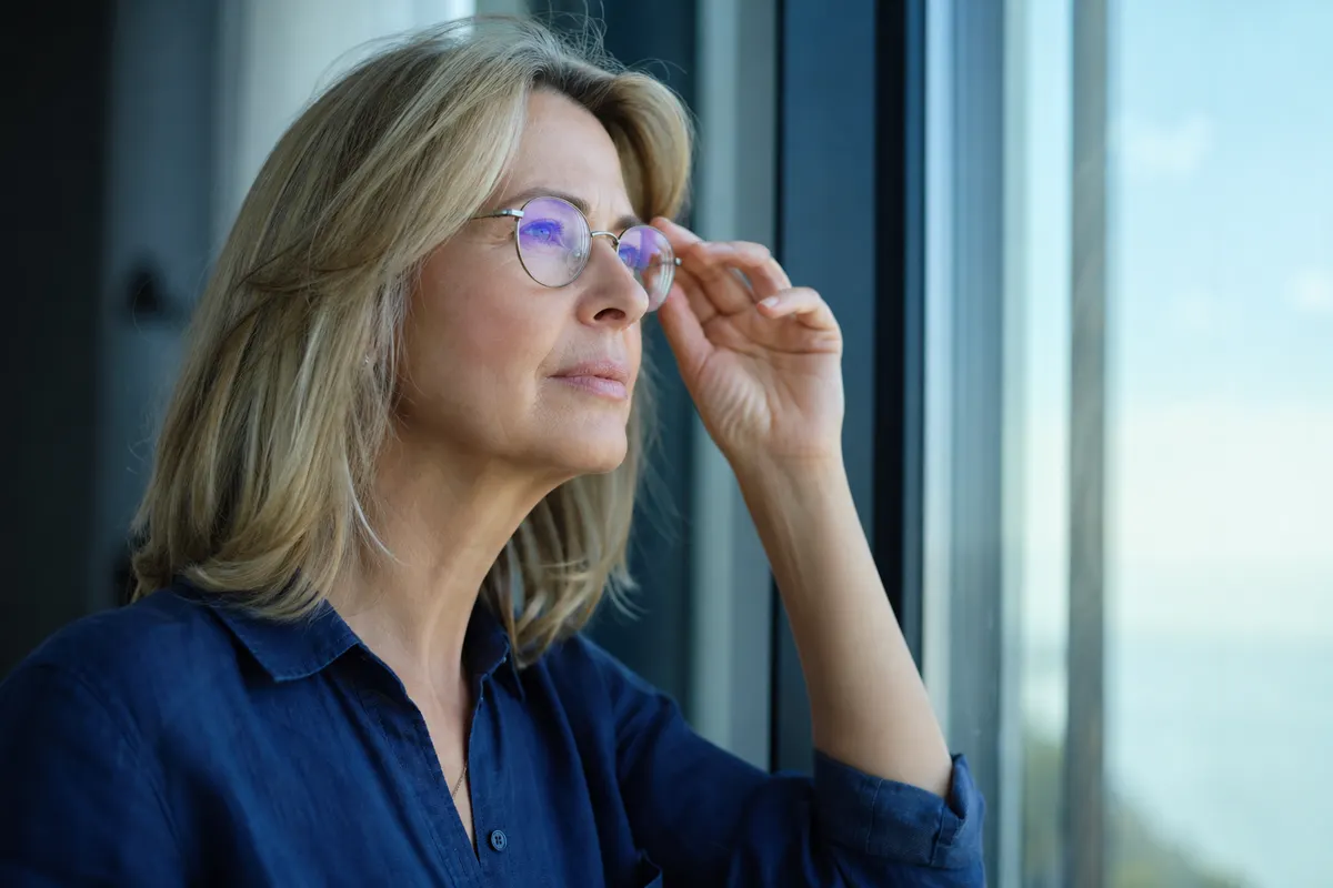 Femme aux cheveux blonds, lunettes à monture fine, tenant la branche de sa monture en regardant un ciel lumineux par une grande baie vitrée.
