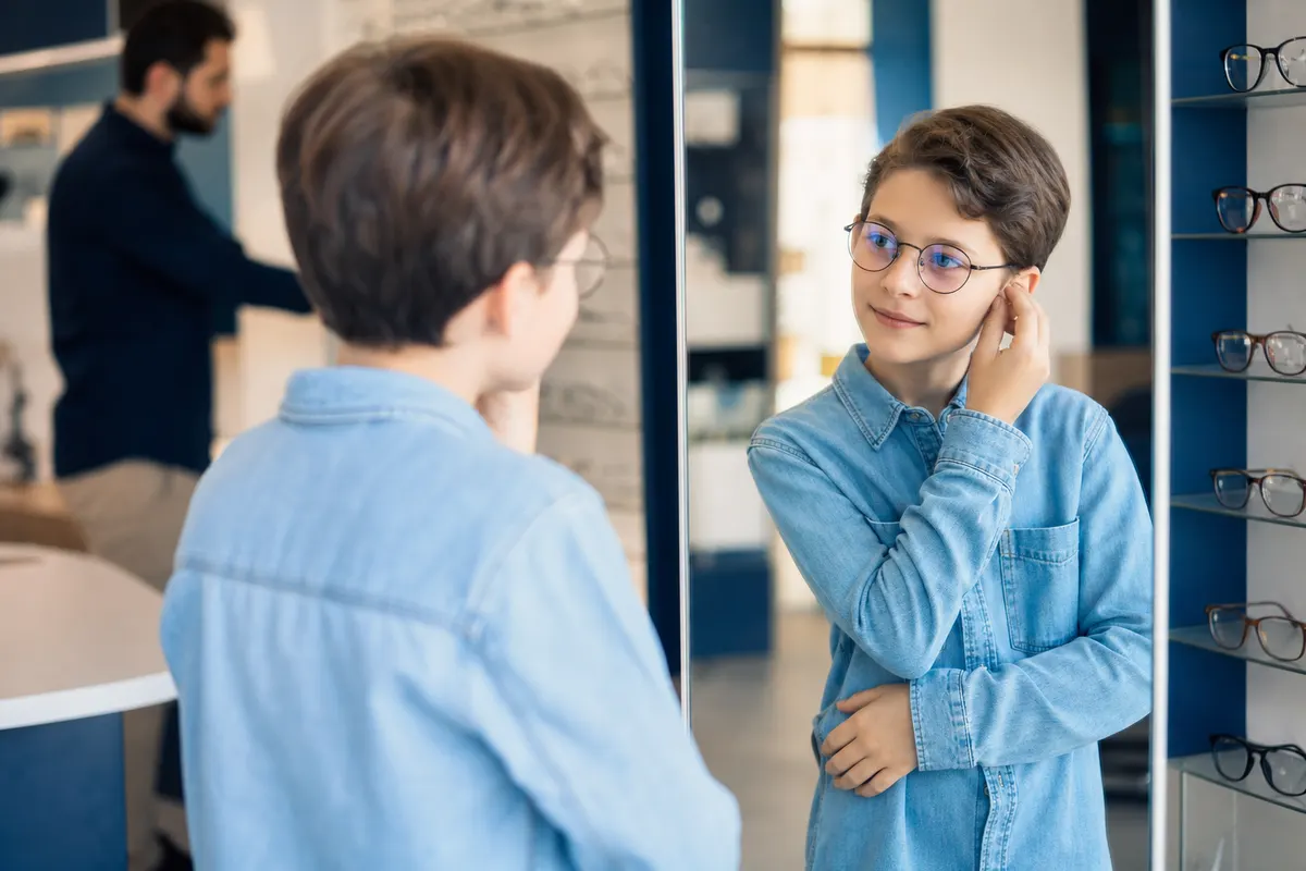 Enfant essayant une paire de lunettes de vue chez l’opticien, comptoir avec étui à lentilles flou en arrière-plan.