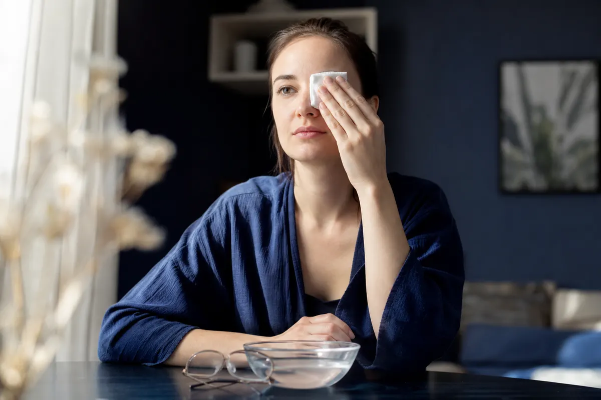 Jeune femme tenant une compresse blanche sur un œil, assise à une table où sont posés une paire de lunettes de vue et un bol d'eau claire.