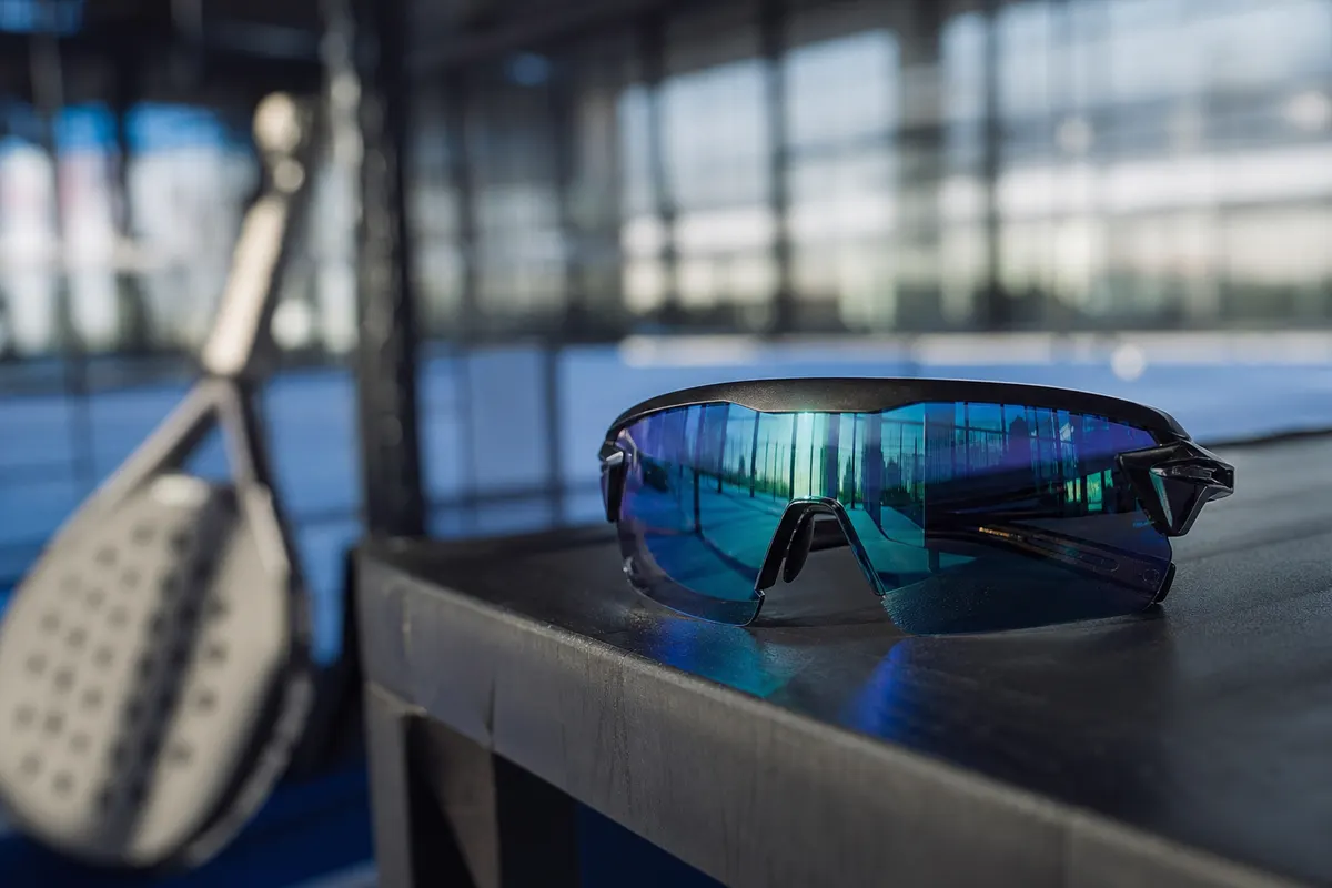 Lunettes de protection sportives enveloppantes posées sur un banc au bord d'un court de padel, lumière naturelle douce, aucune personne visible.