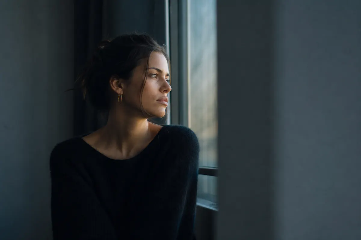 Portrait discret en lumière naturelle douce près d'une fenêtre, regard tourné vers l'extérieur, aucune cigarette ni texte visible.