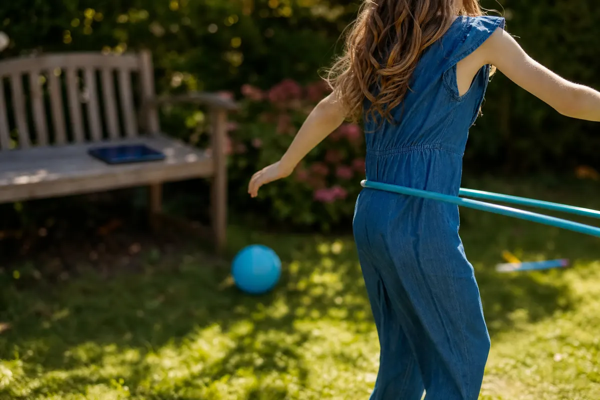 Enfant jouant dehors dans un jardin ensoleillé, tablette posée fermée sur un banc en arrière-plan flou.