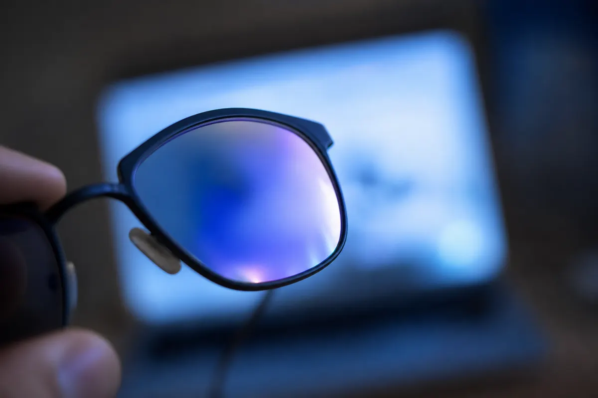 Gros plan sur un verre de lunettes affichant un reflet bleuté caractéristique d'un traitement de surface, tenu face à un écran flou en arrière-plan.