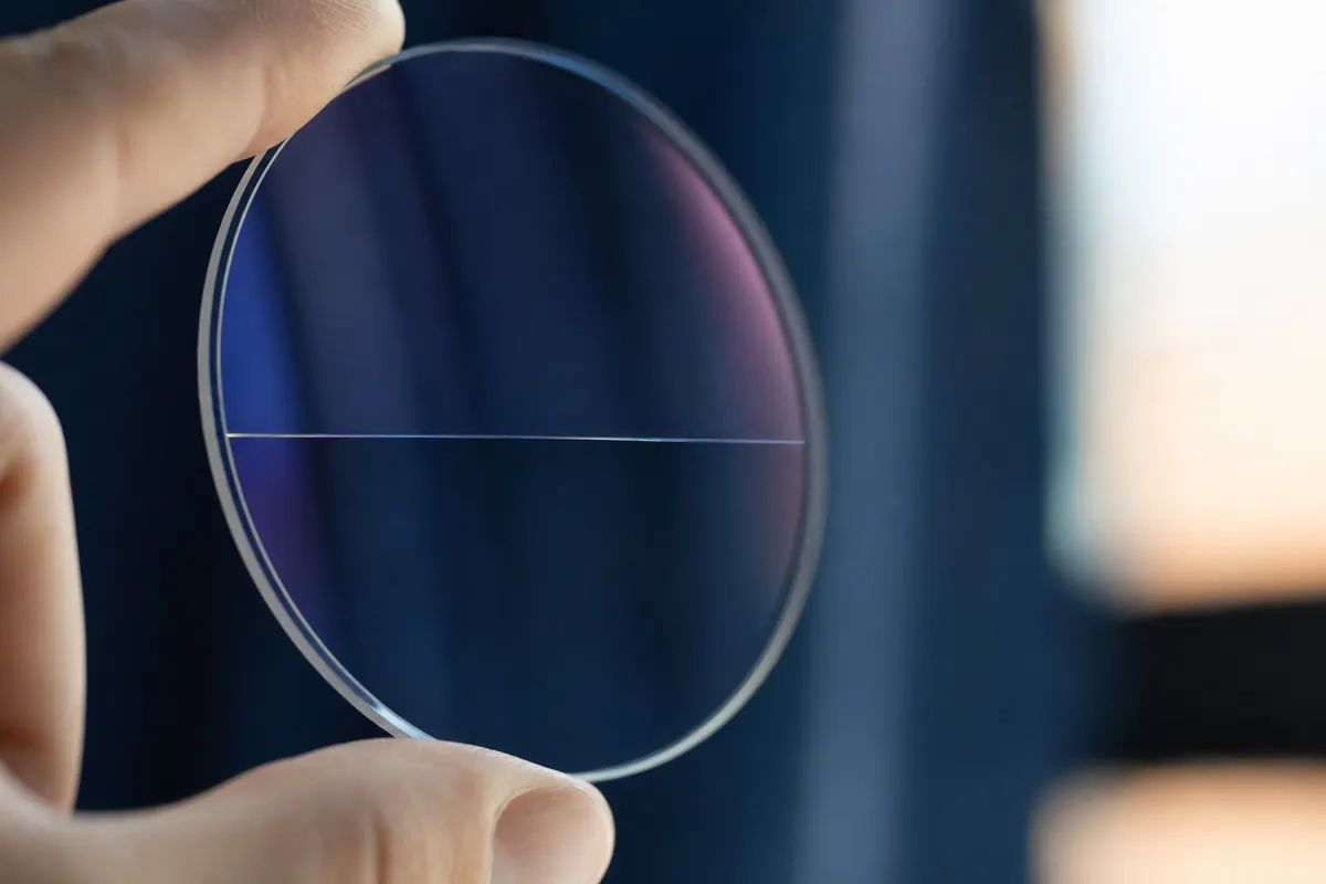 Verre correcteur tenu à contre-jour laissant deviner une ligne de démarcation horizontale, sur fond bleu profond.