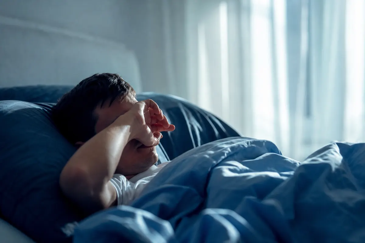 Personne se réveillant dans un lit, se frottant doucement les yeux à la lumière douce du matin, aucun visage entièrement visible.