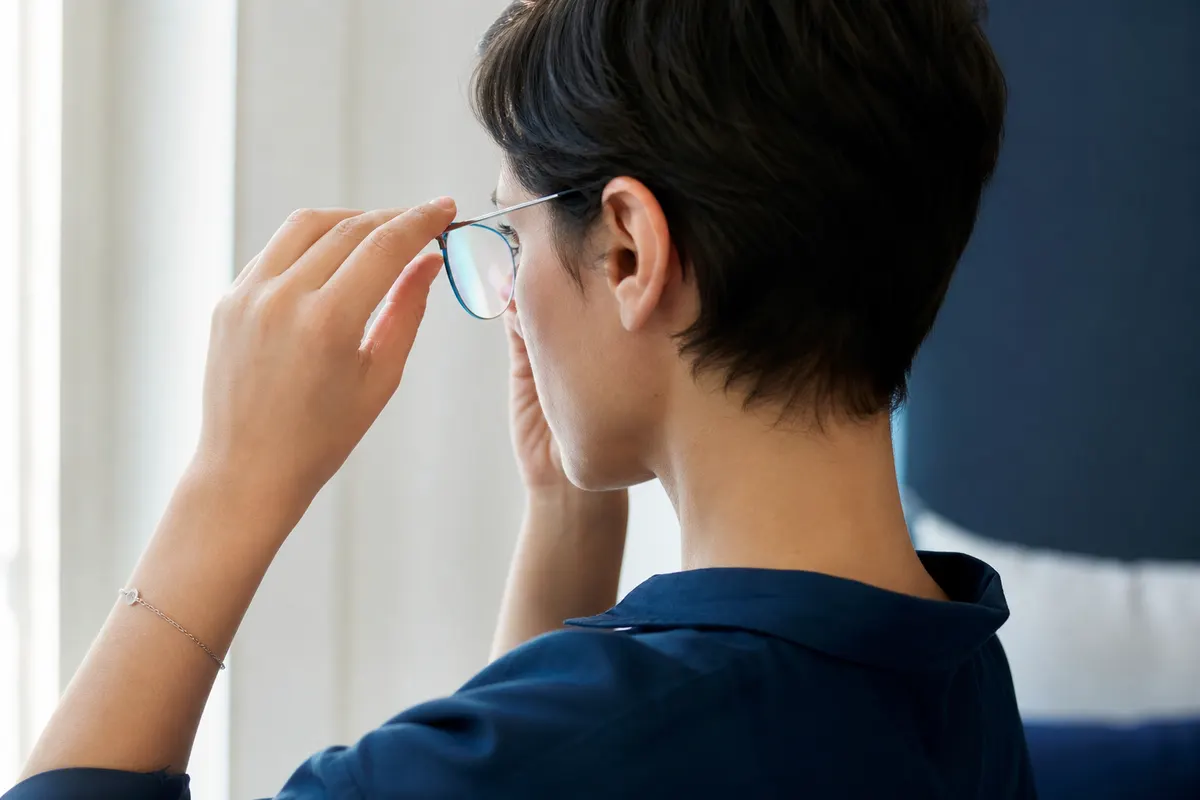 Portrait de trois quarts d'une personne ajustant une paire de lunettes de vue à monture enveloppante, lumière naturelle douce, aucun autre détail visible.